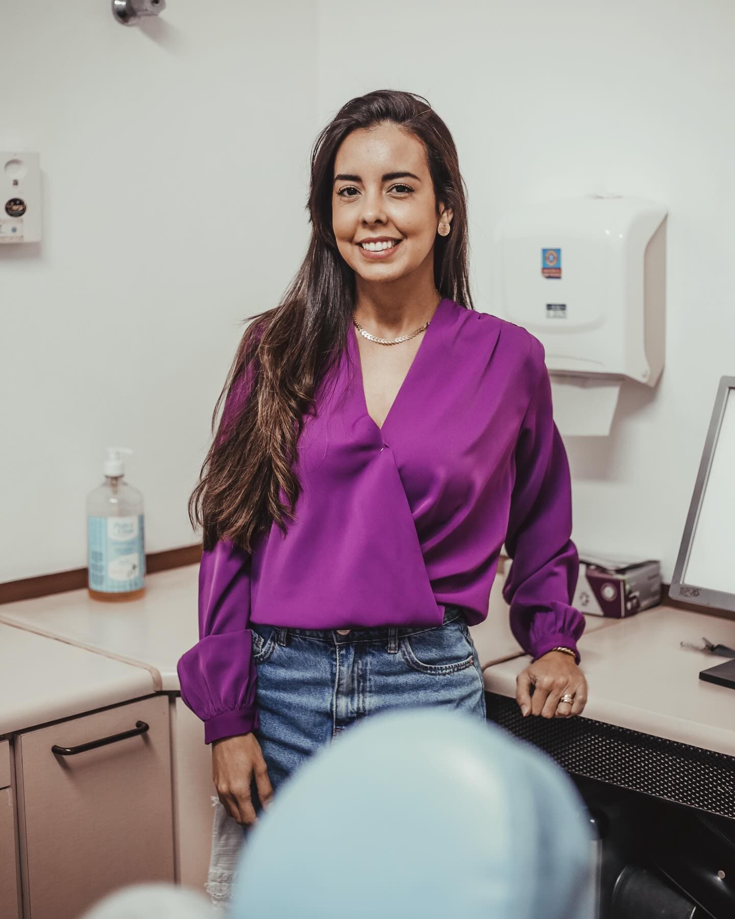 Dra. Marliéria Martins de blusa roxa, sorrindo, encostada na bancada do consultório